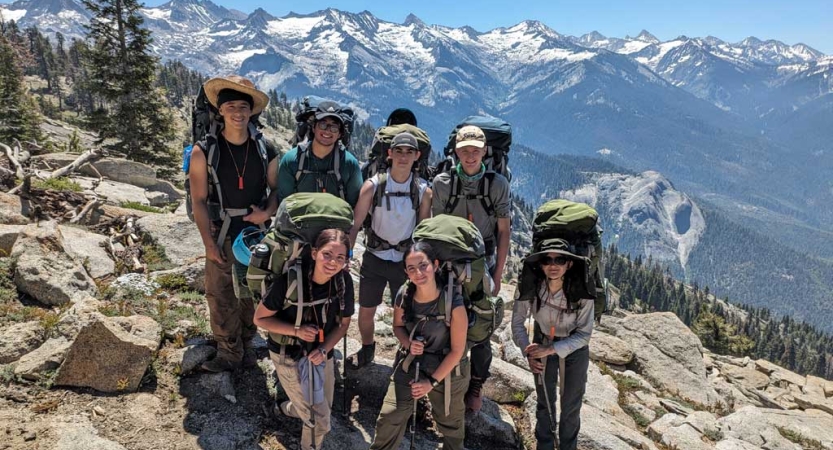 A group of people wearing backpacks stand on rocky ground, high above a snow covered mountainous landscape. 